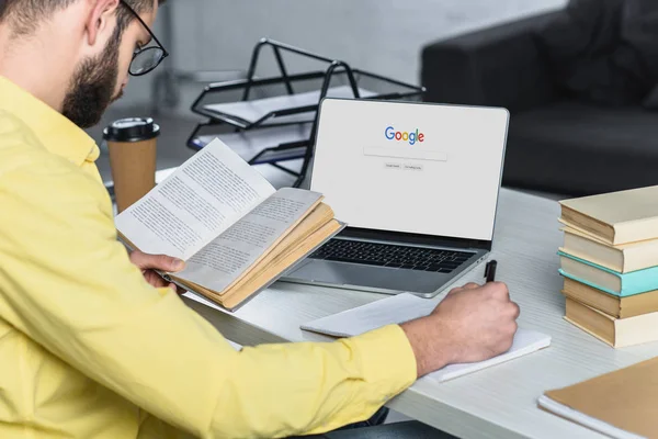 Uomo barbuto che studia con libro vicino computer portatile con google browser sullo schermo in ufficio moderno — Foto stock