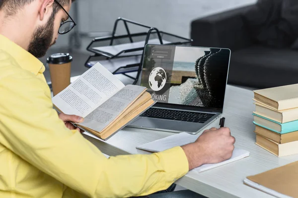 Barbudo hombre estudiando con libro cerca de la computadora portátil con sitio web de reserva en la pantalla en la oficina moderna - foto de stock