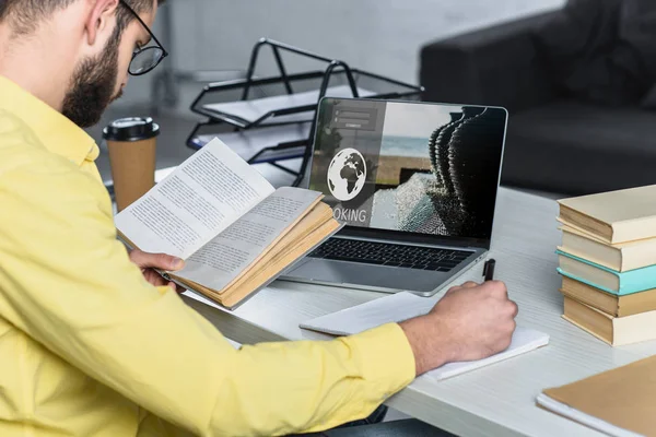 Bearded man studying with book near laptop with booking website on screen in modern office — Stock Photo