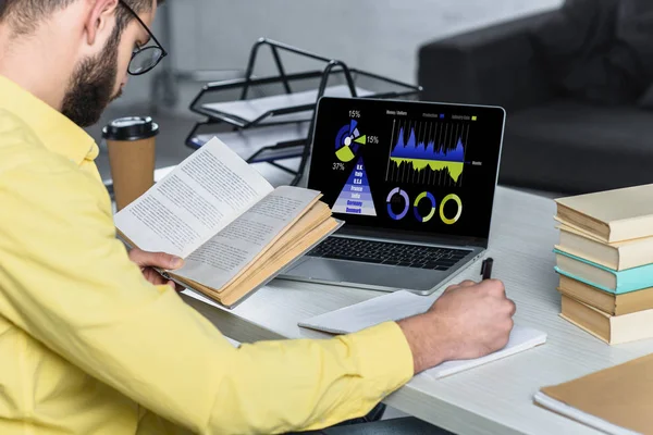 Homme barbu étudiant avec livre près d'un ordinateur portable avec des graphiques et des graphiques à l'écran dans le bureau moderne — Photo de stock