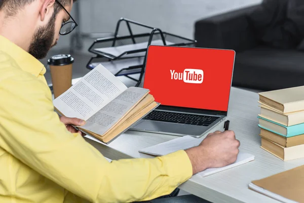 Homem barbudo estudando com livro perto de laptop com site do youtube na tela no escritório moderno — Fotografia de Stock