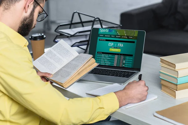Barbudo hombre estudiando con libro cerca de portátil con sitio web sportsbet en la pantalla en la oficina moderna - foto de stock