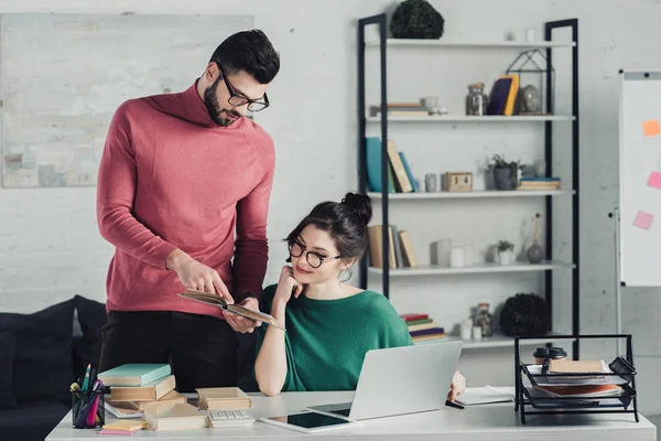 Donna attraente in occhiali che guardano il libro in mani di collega bello in ufficio moderno — Foto stock