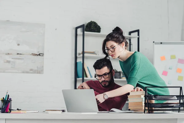 Donna attraente che punta con il dito al computer portatile vicino all'uomo in ufficio moderno — Foto stock