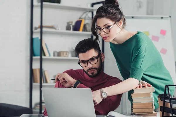 Attraktive Frau mit Brille zeigt mit dem Finger auf Laptop neben Mann in modernem Büro — Stockfoto