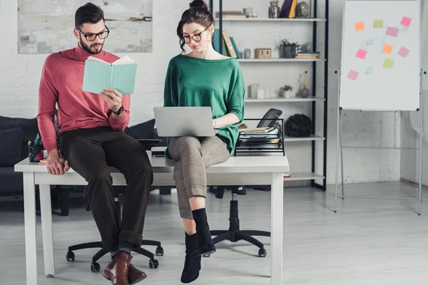 Bärtiger Mann studiert mit Buch, während er auf einem Tisch neben einer Frau mit Laptop sitzt — Stockfoto