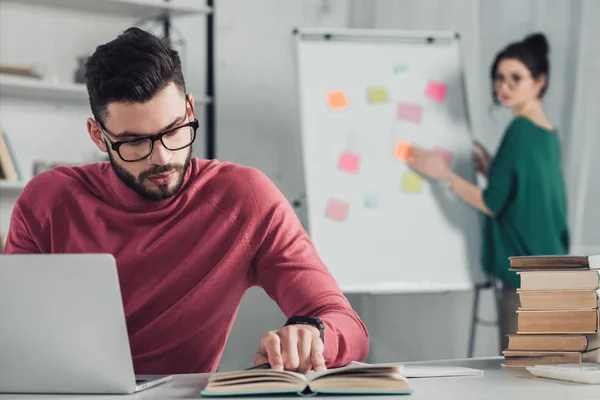 Selective focus of handsome bearded man reading book with woman on background — Stock Photo