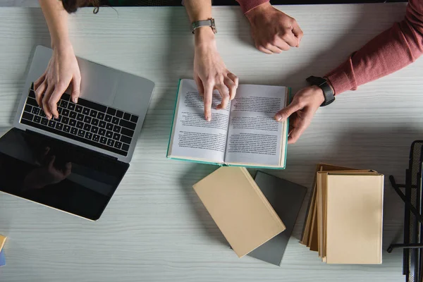 Vista superior de la computadora portátil con pantalla en blanco cerca de la mujer que señala con el dedo en el libro cerca del hombre - foto de stock