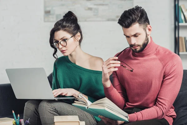 Приваблива жінка в окулярах використовує ноутбук біля бородатого чоловіка, який навчається з книгою — стокове фото
