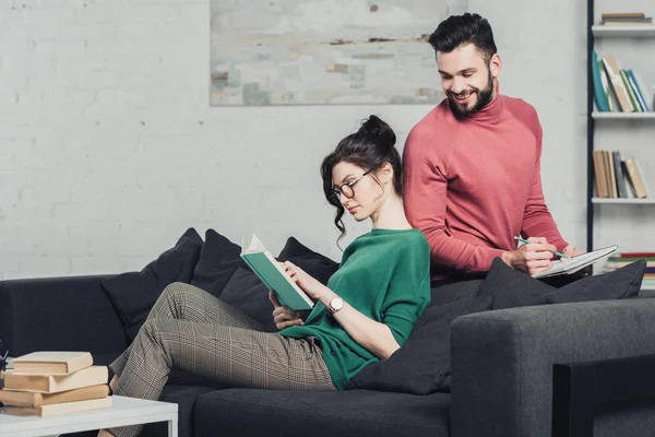 Donna attraente che studia con libro vicino a uomo allegro con notebook e matita in mano — Foto stock