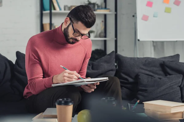 Bel homme en lunettes écrivant dans un cahier tout en étudiant à la maison — Photo de stock