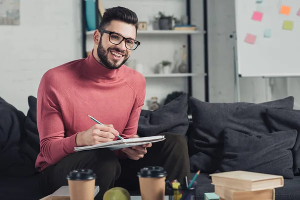 Fröhlicher Mann mit Brille, der zu Hause in Notizbuch schreibt — Stockfoto