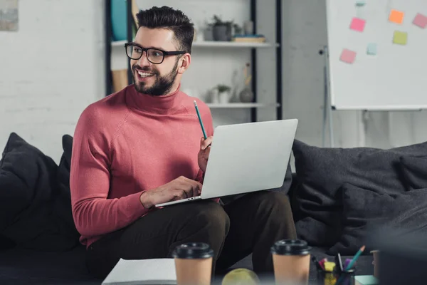 Cheerful bearded man in glasses using laptop while sitting on sofa — Stock Photo