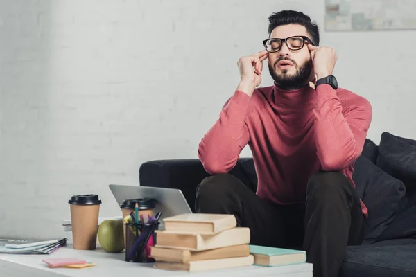 Hombre barbudo en gafas sentado cerca del ordenador portátil en el sofá - foto de stock