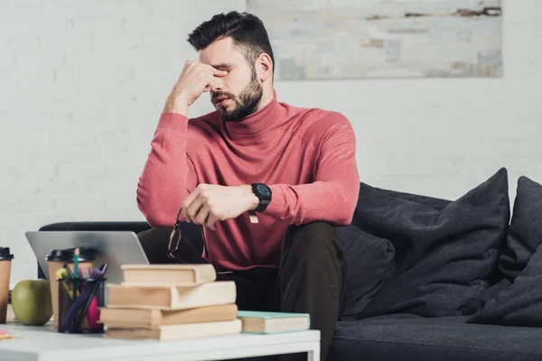 Hombre cansado sosteniendo gafas y sentado en el sofá cerca de la computadora portátil y libros — Stock Photo