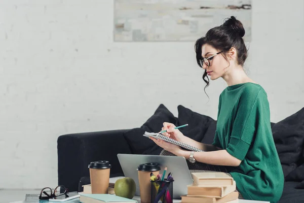 Femme attrayante dans des lunettes écriture dans un cahier tout en étudiant à la maison — Photo de stock