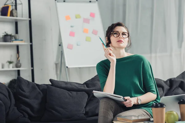 Pensive woman in glasses holding notebook and sitting on sofa at home — Stock Photo