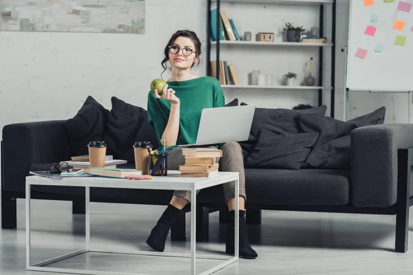 Atractiva mujer en gafas sosteniendo manzana mientras está sentado con el ordenador portátil en el sofá - foto de stock