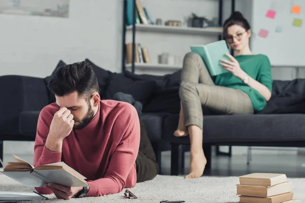 Messa a fuoco selettiva dell'uomo stanco che studia con il libro mentre giace sul tappeto con la donna sullo sfondo — Foto stock