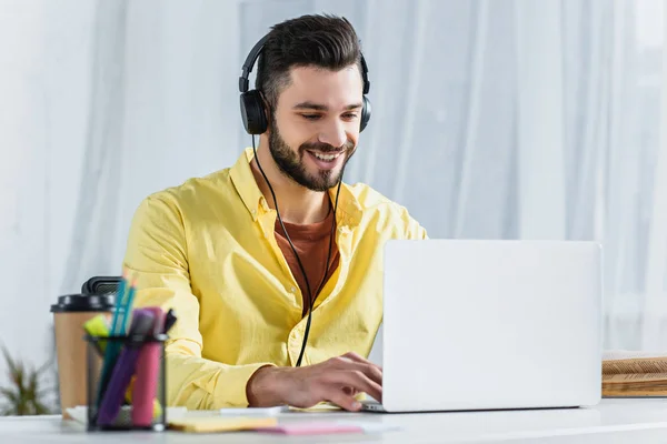 Homem alegre bonito em fones de ouvido usando laptop — Fotografia de Stock