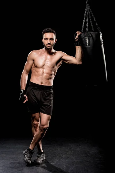 Schöner Boxer, der mit überkreuzten Beinen neben Boxsack auf schwarzem Hintergrund steht — Stockfoto