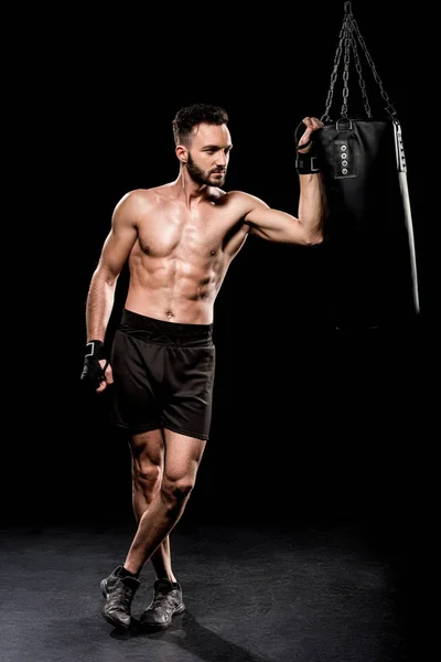 Kurzloser Boxer, der mit überkreuzten Beinen neben Boxsack auf schwarzem Hintergrund steht — Stockfoto