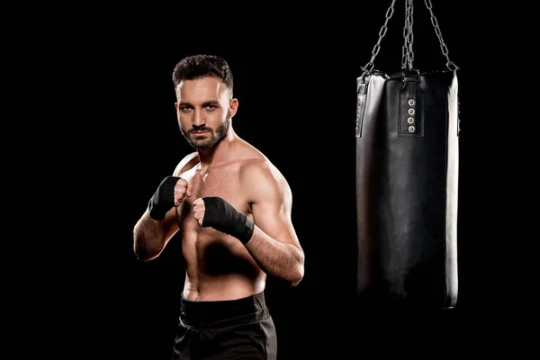 Shortless man looking at camera near punching bag isolated on black — Stock Photo