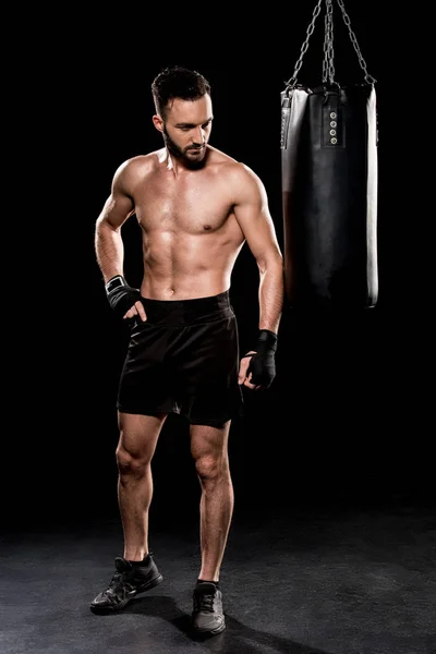 Shortless bearded man looking at punching bag on black background — Stock Photo