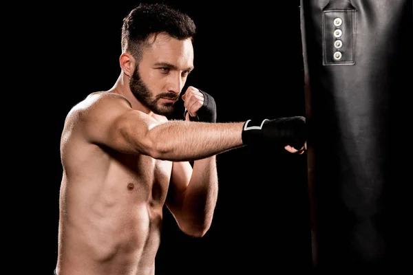 Shortless bearded man hitting punching bag isolated on black — Stock Photo