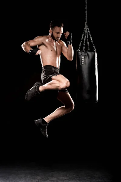 Handsome boxer performing flying kick near punching bag on black background — Stock Photo