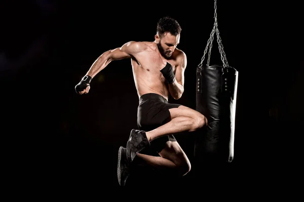 Boxeador sin cortocircuito realizando patada voladora cerca de saco de boxeo aislado en negro - foto de stock