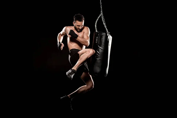 Shortless man performing flying kick near punching bag isolated on black — Stock Photo