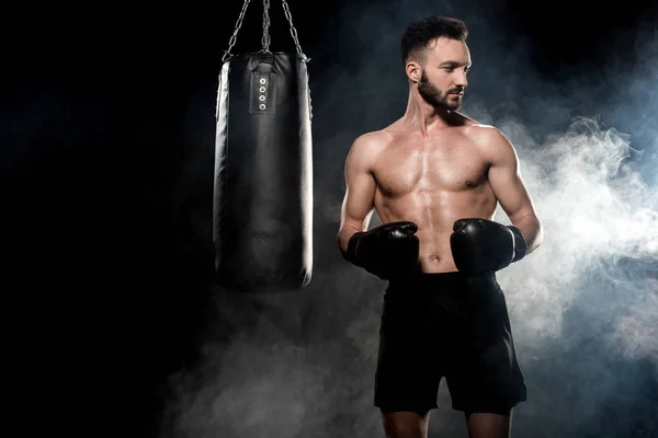 Pensive sportsman in boxing gloves standing near punching bag on black with smoke — Stock Photo