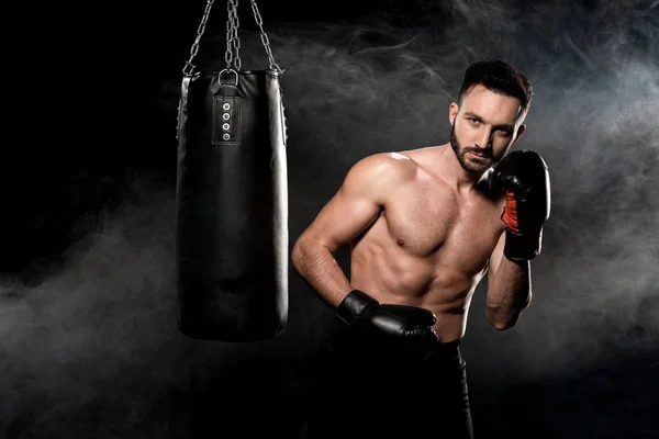 Handsome sportsman in boxing gloves standing near punching bag on black with smoke — Stock Photo