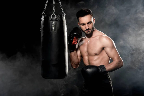 Muscular sportsman in boxing gloves standing near punching bag on black with smoke — Stock Photo