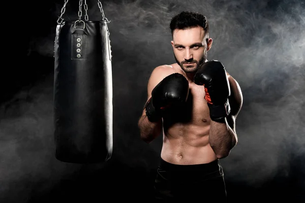 Muscular athlete standing in boxing pose near punching bag on black with smoke — Stock Photo