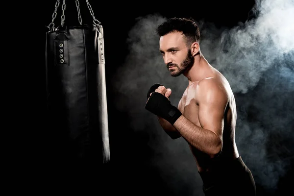 Atleta muscular de pie en pose de boxeo y mirando el saco de boxeo en negro con humo - foto de stock