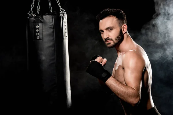 Muskulöser Mann steht in Boxerpose und blickt in die Kamera neben Boxsack auf schwarz mit Rauch — Stockfoto