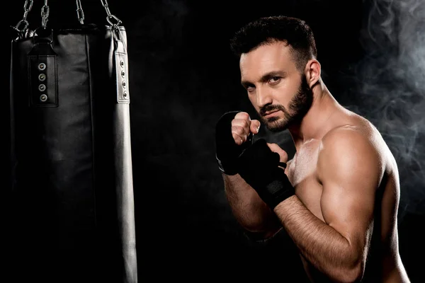 Combattant musculaire debout dans la posture de boxe près d'un sac de boxe isolé sur noir — Photo de stock