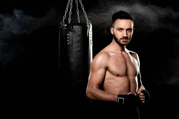 Athletic handsome man standing near punching bag on black with smoke — Stock Photo