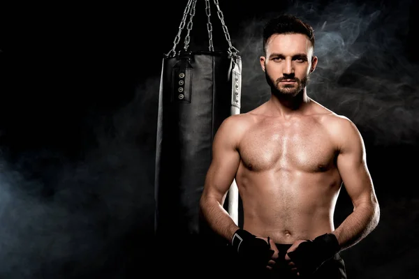 Handsome shortless boxer standing near punching bag on black with smoke — Stock Photo