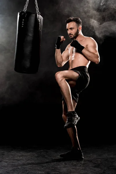 Handsome man exercising near punching bag on black with smoke — Stock Photo