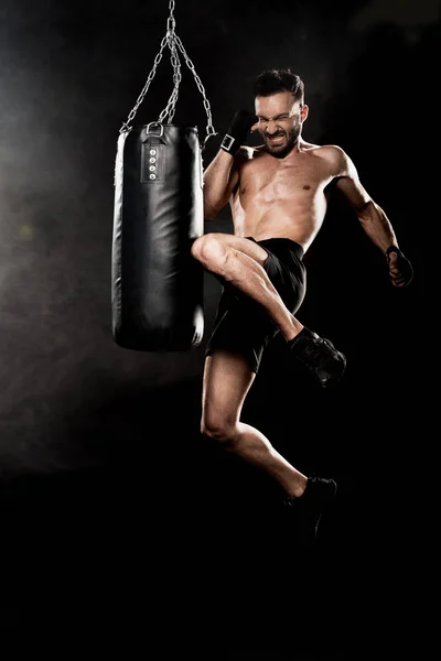 Athletic boxer performing flying kick near punching bag on black with smoke — Stock Photo