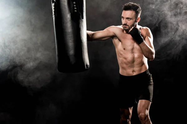 Handsome athletic boxer exercising with punching bag on black with smoke — Stock Photo