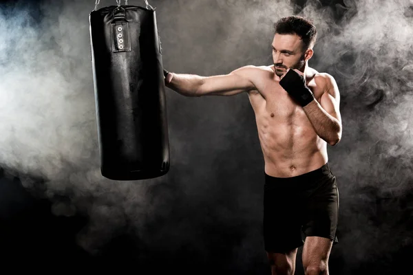 Shortless athletic boxer exercising with punching bag on black with smoke — Stock Photo