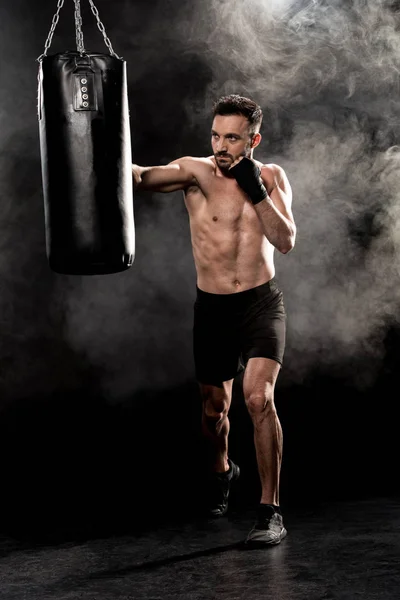 Shortless athletic boxer kicking punching bag on black with smoke — Stock Photo