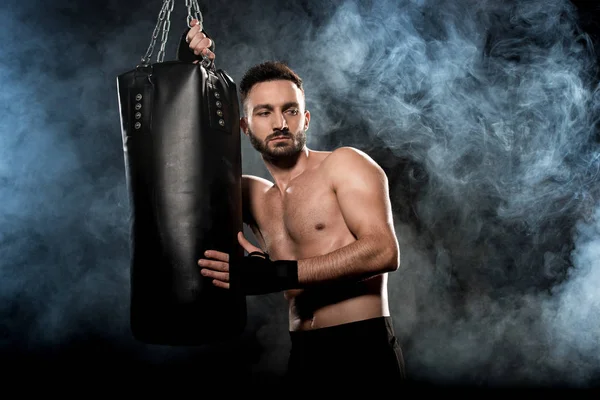 Shortless athletic boxer holding punching bag on black with smoke — Stock Photo