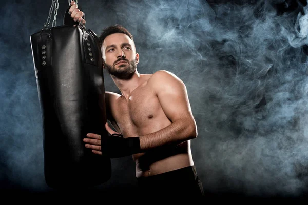 Pensive athletic boxer holding punching bag on black with smoke — Stock Photo