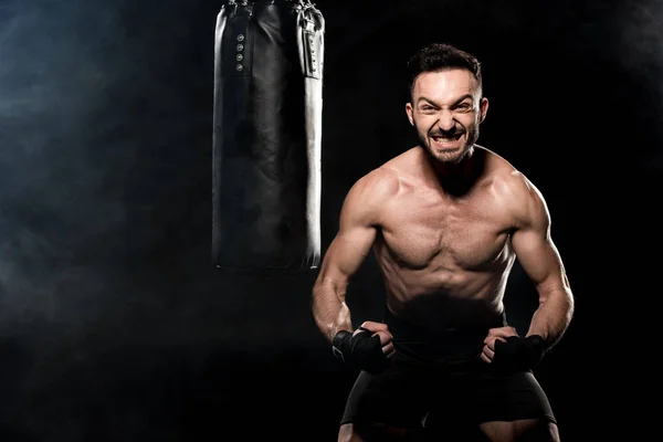 Angry boxer showing muscles punching bag on black with smoke — Stock Photo