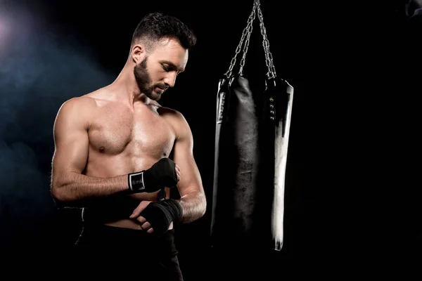 Schöner athletischer Boxer, der Handschuhe in der Nähe eines Boxsacks auf schwarzem Hintergrund einwickelt — Stockfoto